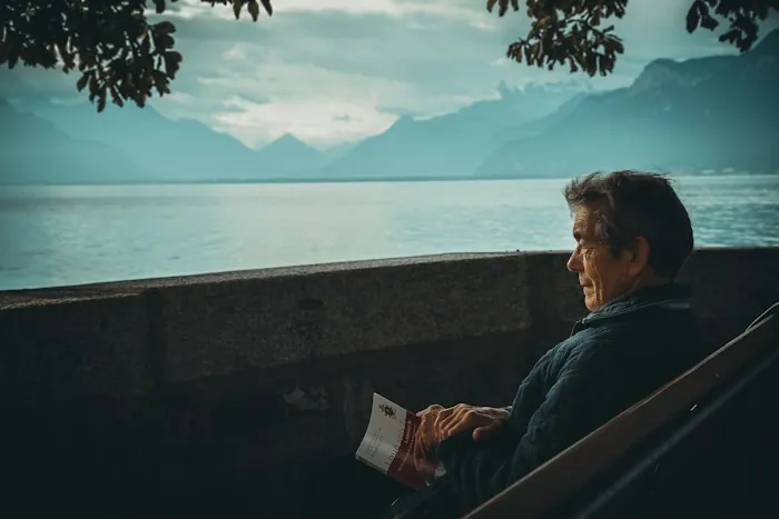 A man sitting by a lake with mountains behind him, holding a book