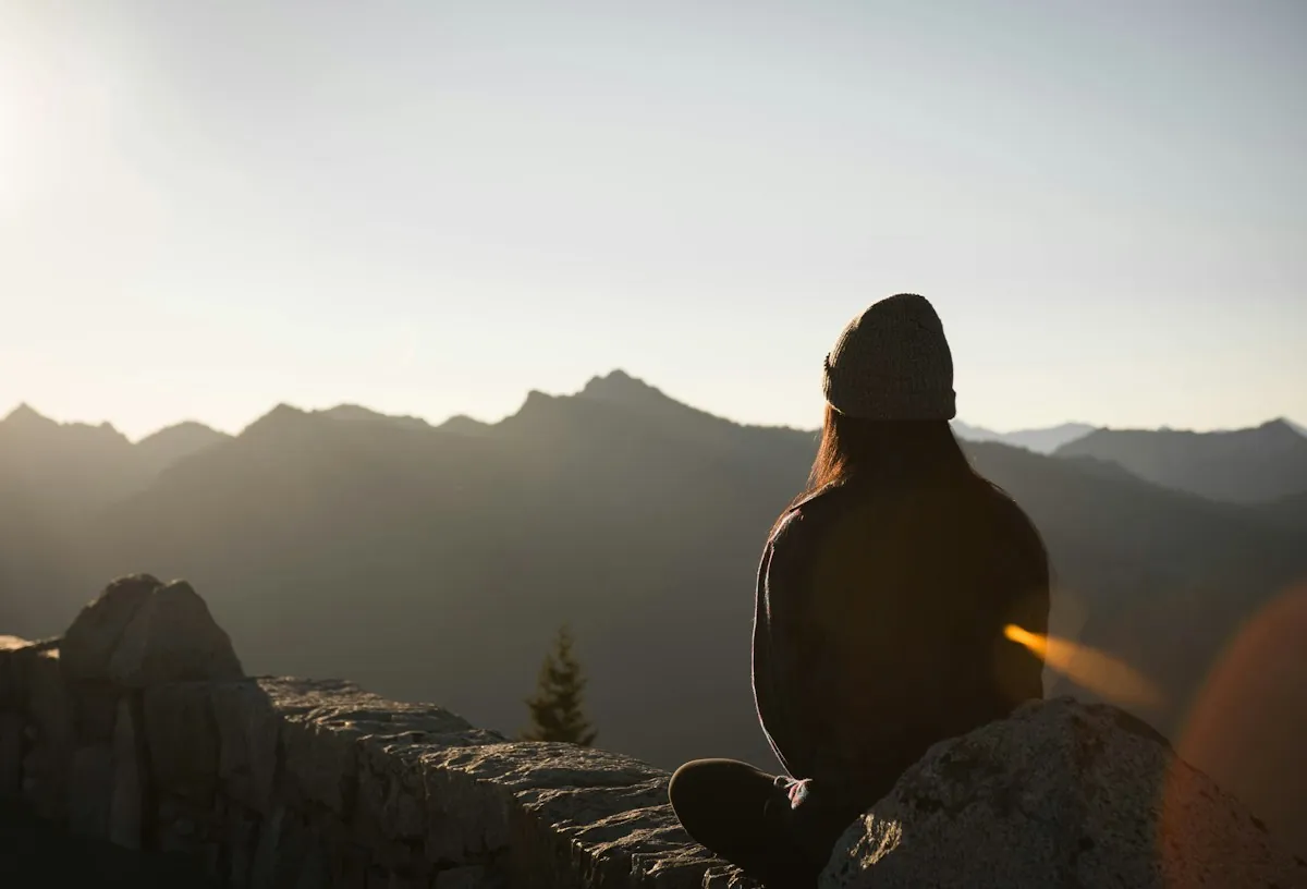 A person in a beanie sitting on a rock, looking out at a mountain range at dusk