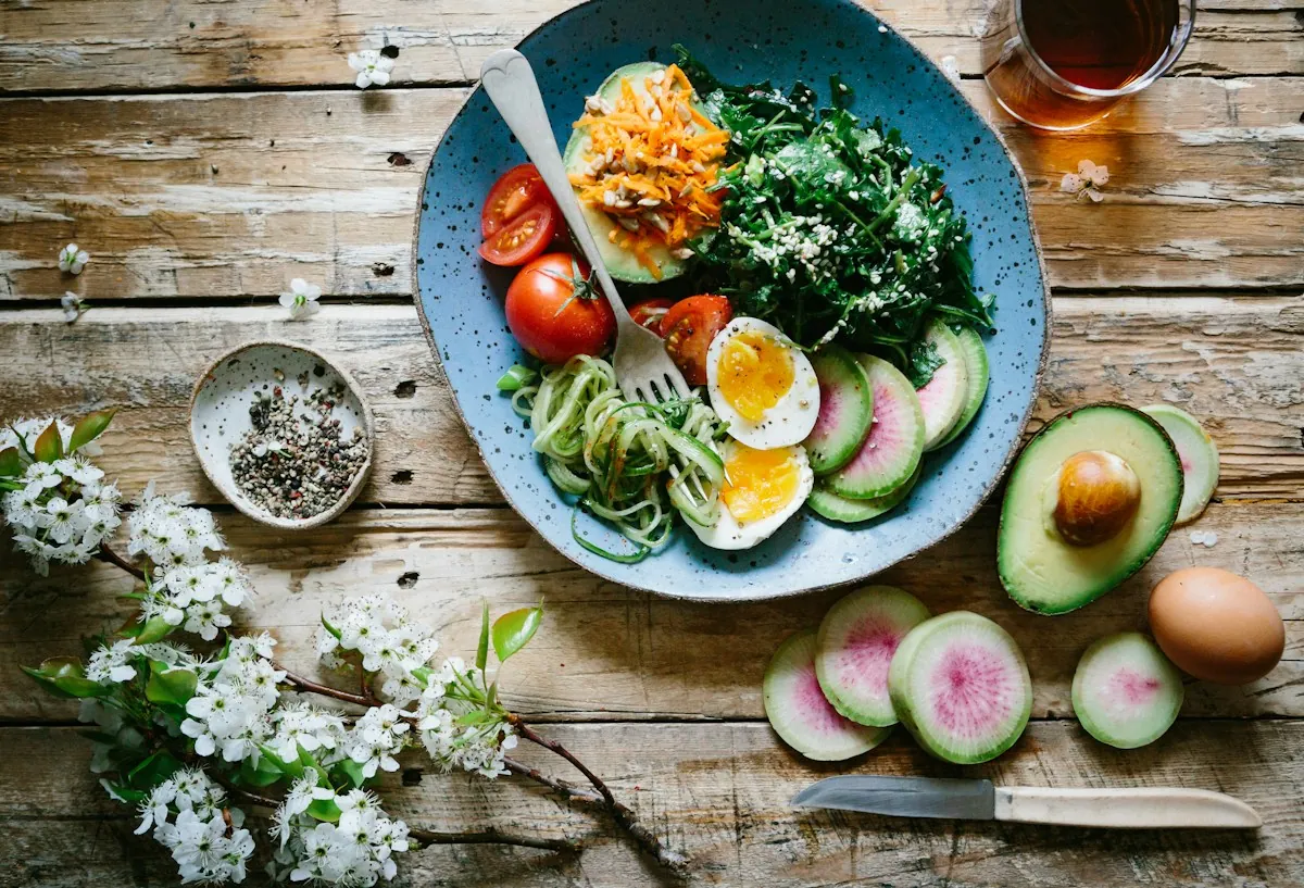 A blue bowl of vegetable noodles, greens, tomatoes and boiled egg beside sliced avocado and radish on a wooden table