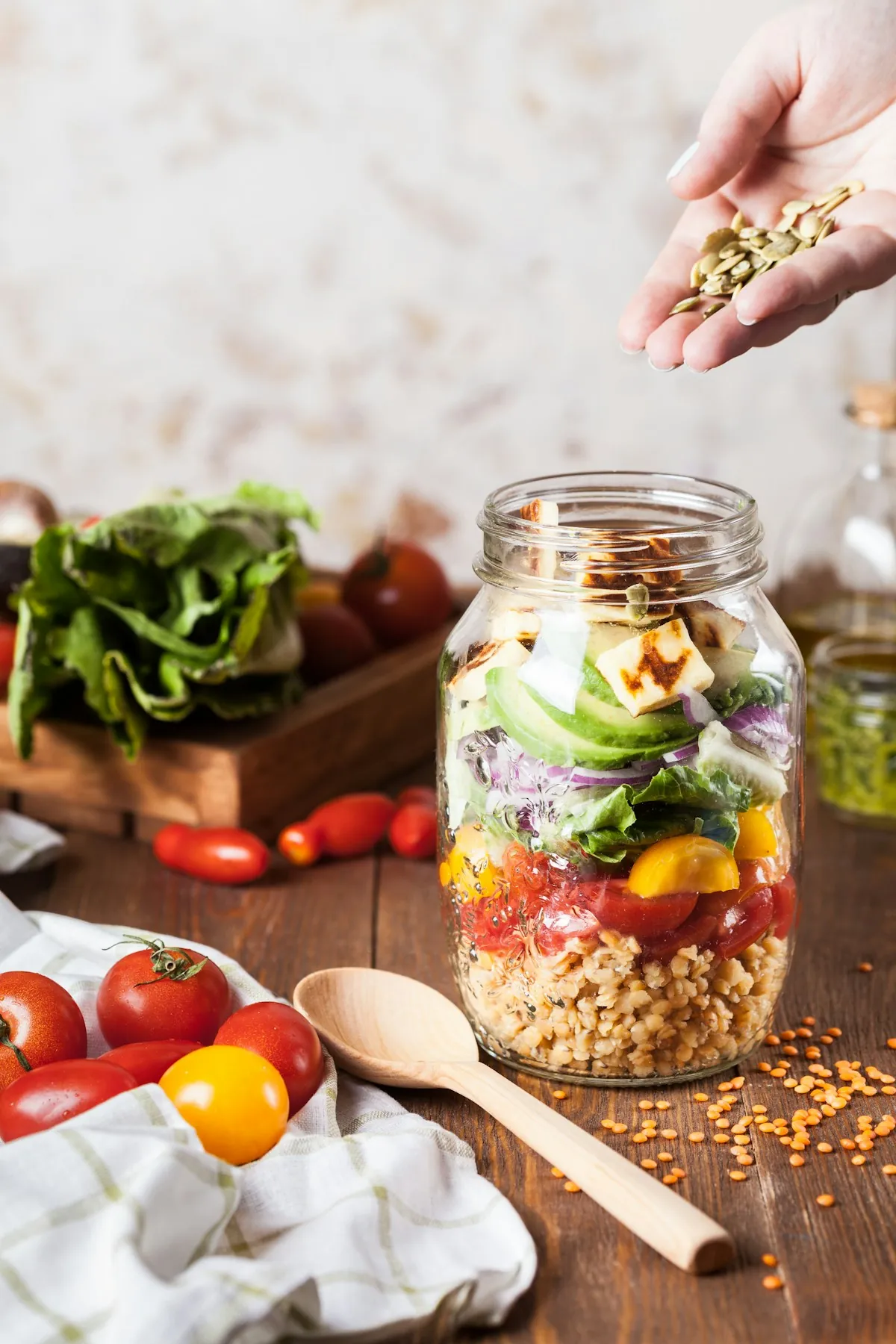 A glass jar layered with grains, avocado and vegetables next to fresh tomatoes and greens