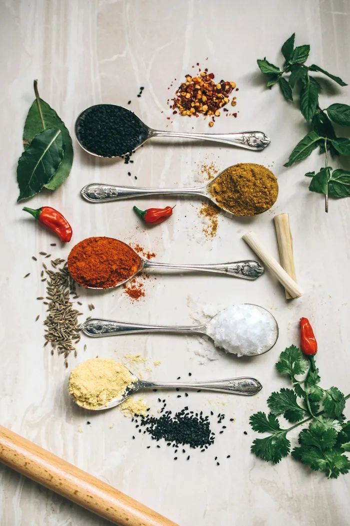 Silver spoons holding colorful spices and salt on a white surface, with chilies and leaves
