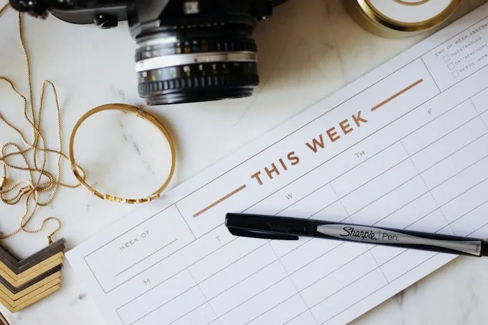 A weekly planner page and pen on a marble desk beside a camera and gold bracelets