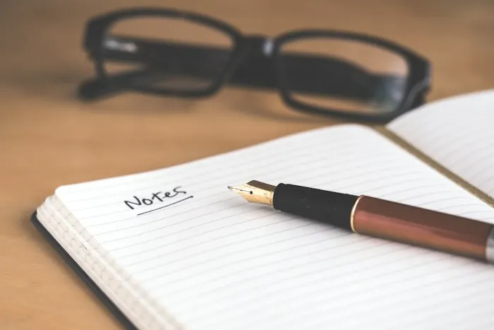 An open notebook with a fountain pen and reading glasses on a wooden desk