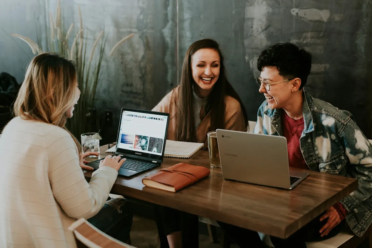 Three coworkers laughing together over laptops at a wooden table