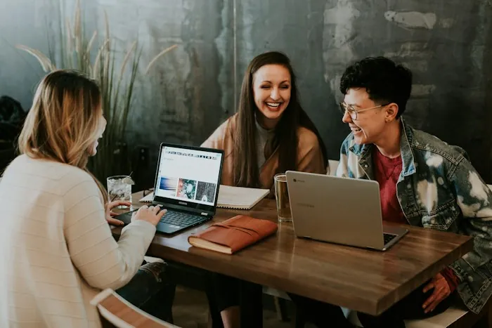 Three coworkers laughing together over laptops at a wooden table