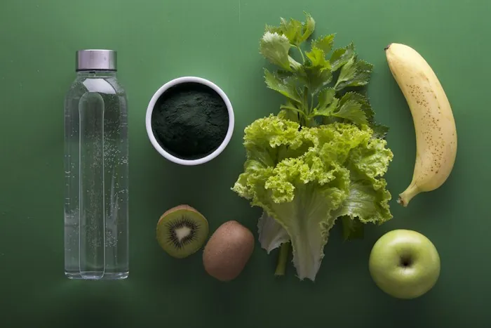 A glass water bottle beside fresh greens, fruit and a bowl of green powder