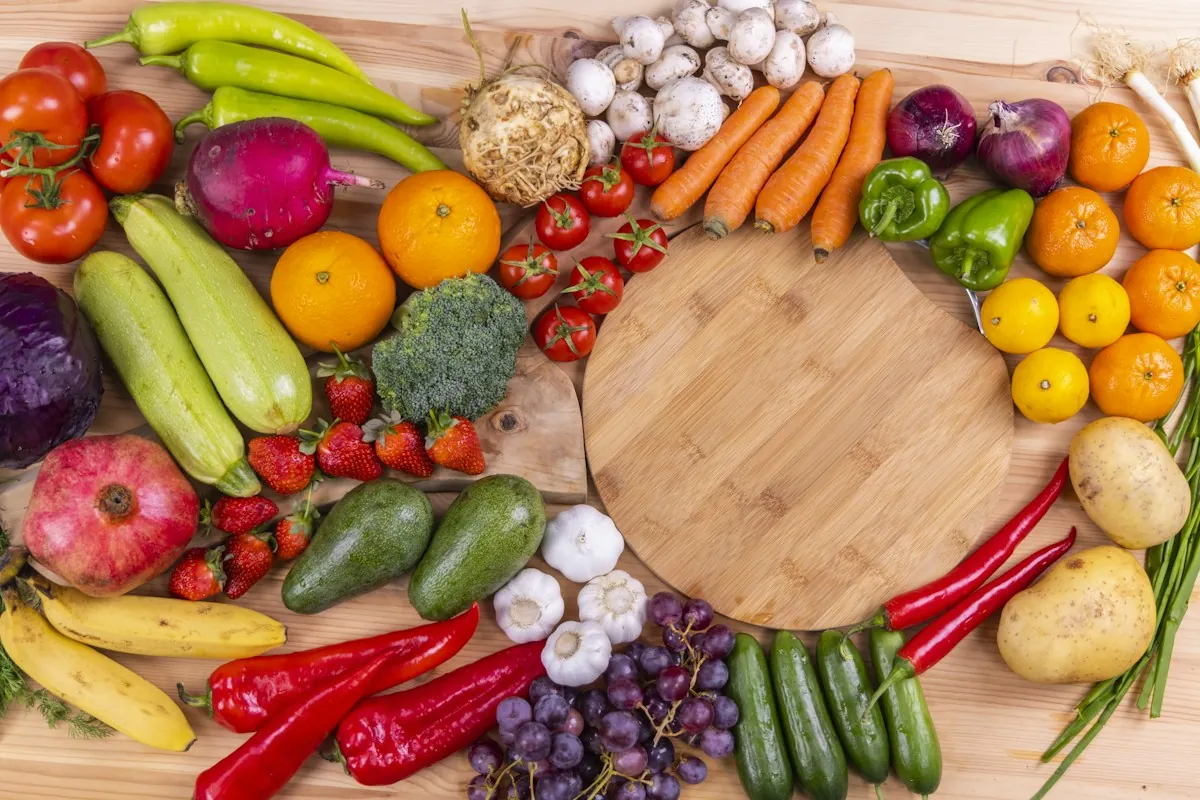 A wooden board surrounded by colorful whole vegetables and fruit, including peppers, carrots, tomatoes, grapes and citrus