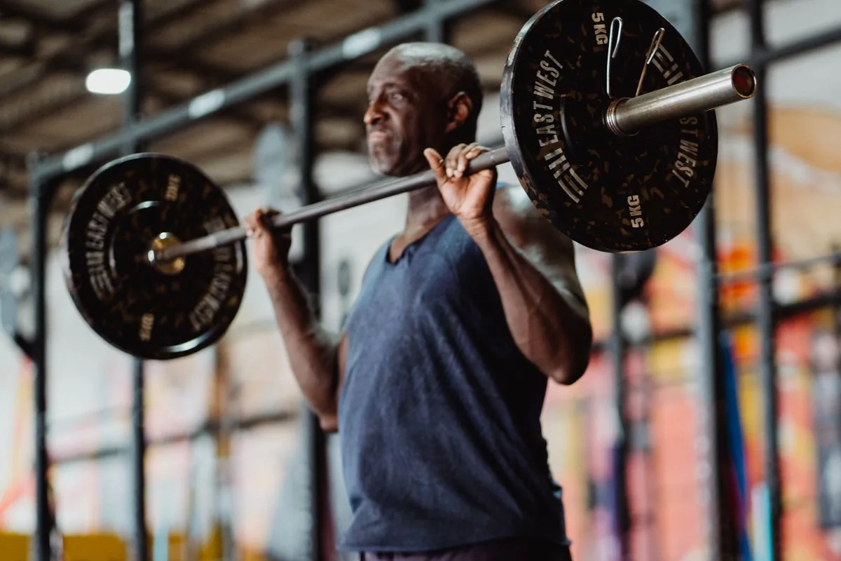 An older man lifting a barbell onto his shoulders in a gym, focused on his form