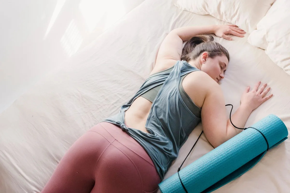 A woman in activewear resting on a bed beside a rolled yoga mat