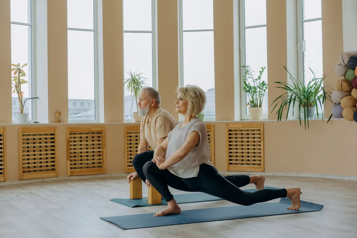 An older couple doing a gentle supported lunge stretch together in a bright room