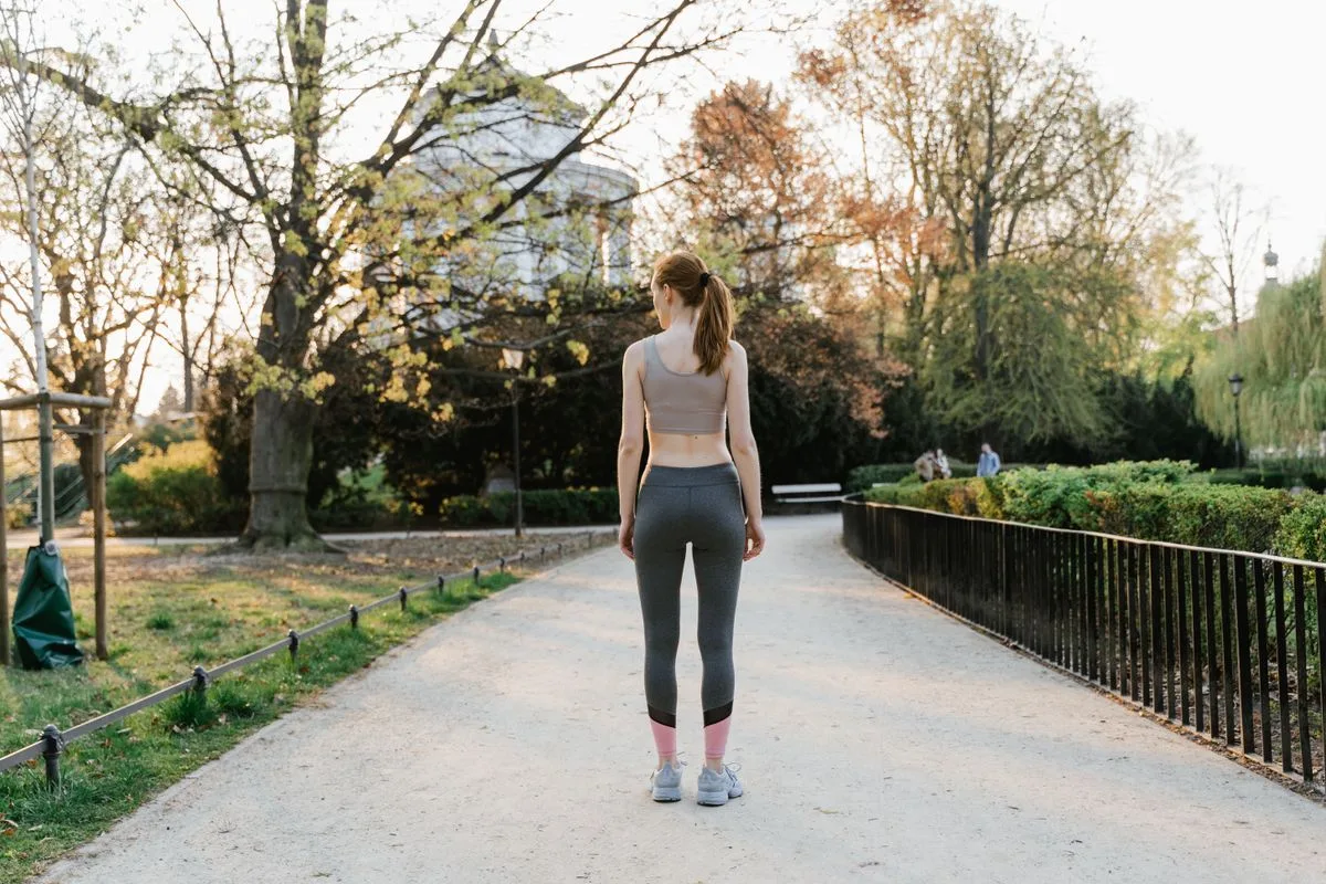 A woman standing on a park path in workout clothes, seen from behind