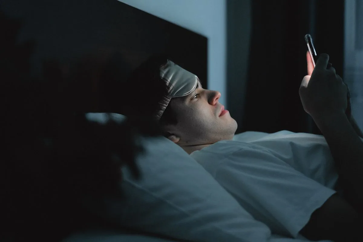 A man wearing a sleep mask pushed up on his forehead, checking his phone late at night