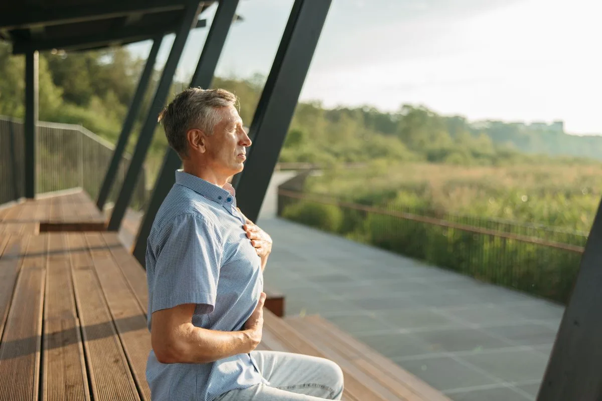 An older man sitting outdoors with his hand on his chest, practicing a calming breath