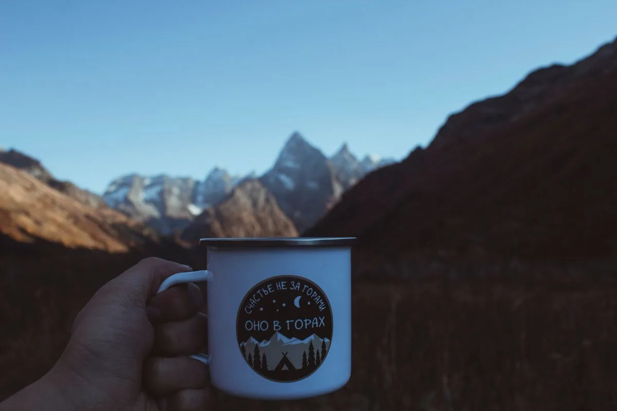 A hand holding a camping mug with mountains in the background, enjoying a quiet moment outdoors
